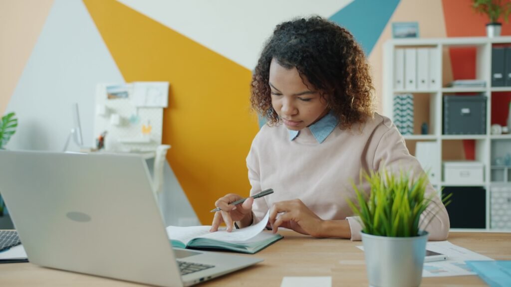 Person writing research notes at a desk with a laptop, representing NotebookLM research workflows